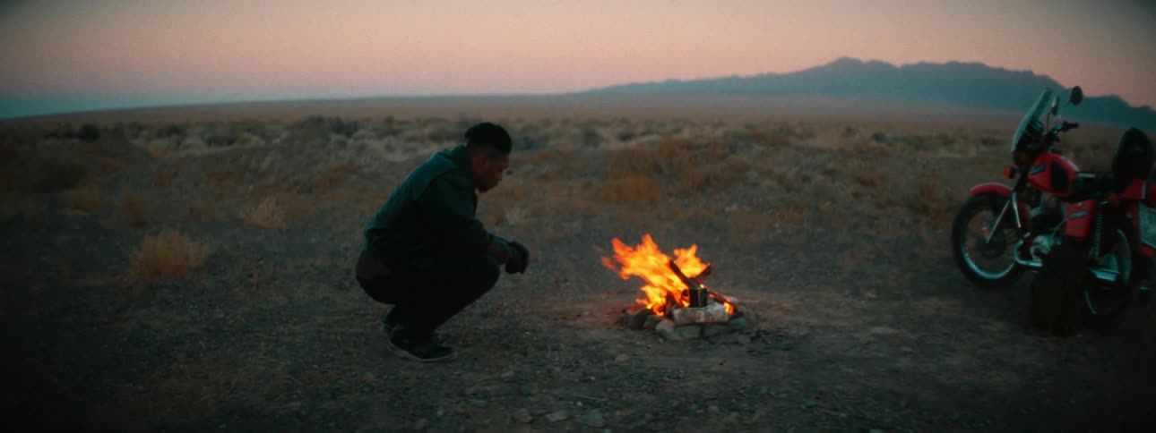 A man on a solo motorcycle trip crouches to warm his hands by a small campfire in the desert at dusk
