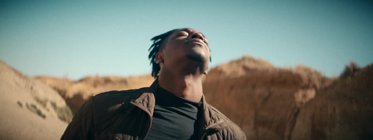 A slow zoom-out reveals a young man with his eyes closed, serenely enjoying the warmth of the sun in a vast desert canyon