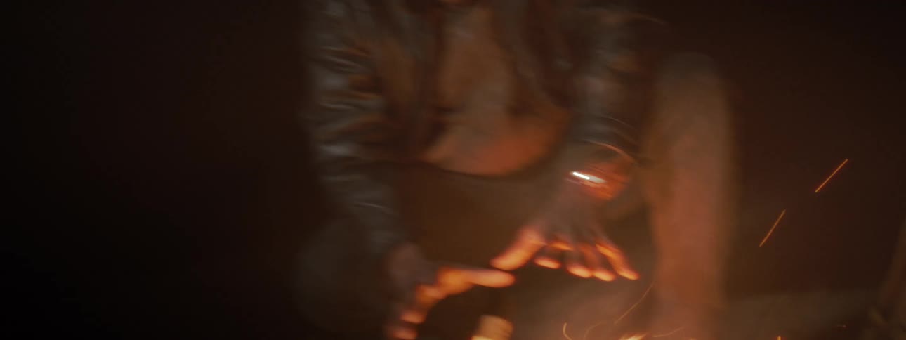 A young man in a leather jacket warms his hands by a crackling campfire in the dark of night