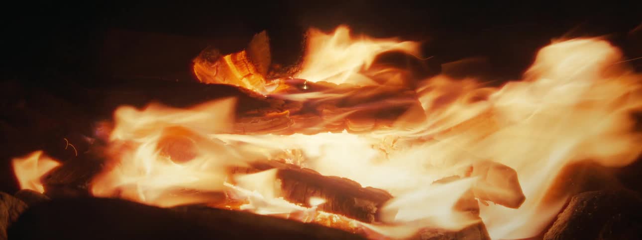 A close-up shot of a roaring fire with bright orange flames engulfing logs in the dark