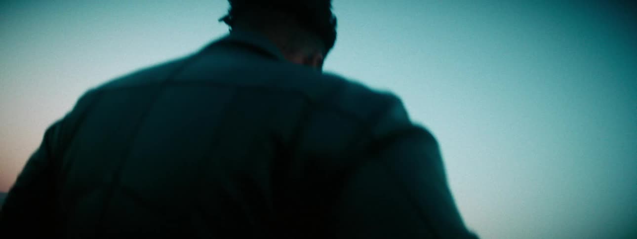 A pensive man stands up in slow motion to look out over a vast desert landscape at dusk