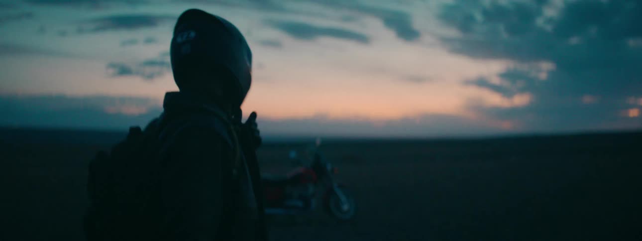 A silhouetted motorcyclist adjusts their helmet and walks toward their bike parked in a vast, open landscape at dusk