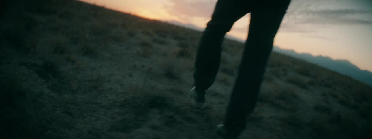 Slow-motion close-up of a person's boots walking up a grassy hill towards the warm glow of the sunset