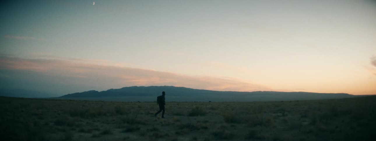 A solitary figure with a backpack journeys through a vast desert at dusk under a crescent moon, with distant mountains silhouetted against the fading light