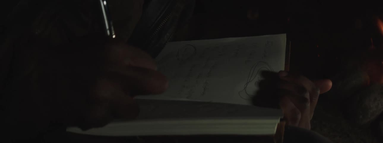 A close-up shot of someone writing in their journal, illuminated by the warm glow of a campfire at night