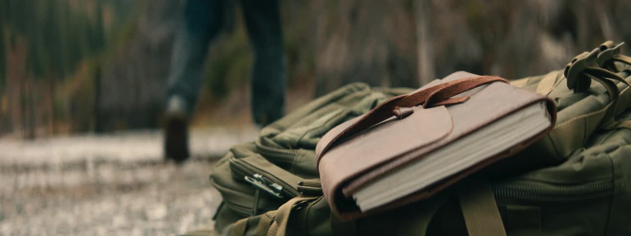 An adventurer's leather-bound journal rests on a backpack as they walk into the wilderness