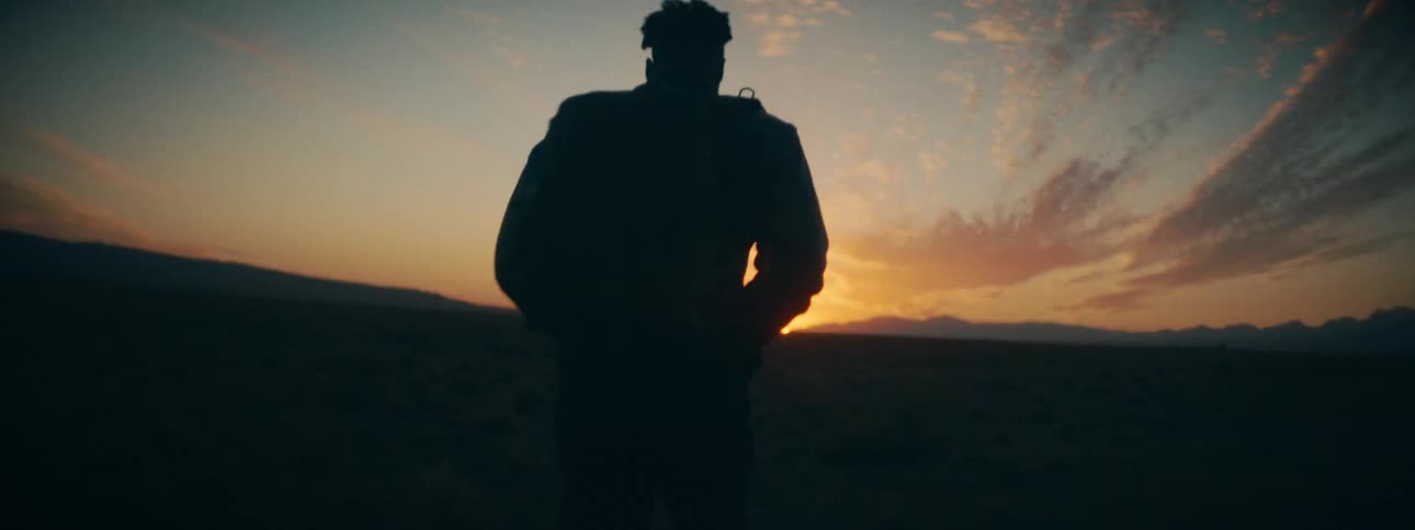 A silhouetted man with dreadlocks walks through a vast, open landscape towards the warm glow of the setting sun