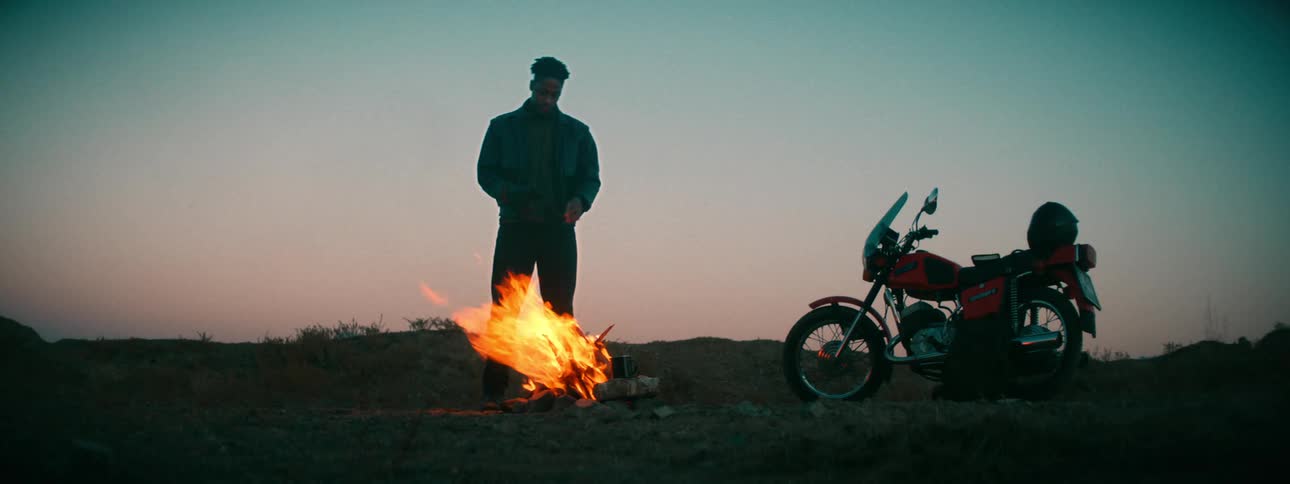 A young man stands next to a crackling campfire at twilight with his motorcycle parked nearby