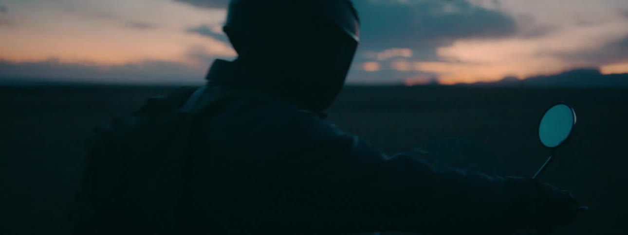 A silhouetted motorcyclist with a backpack gets on their bike in slow motion in an open landscape at dusk