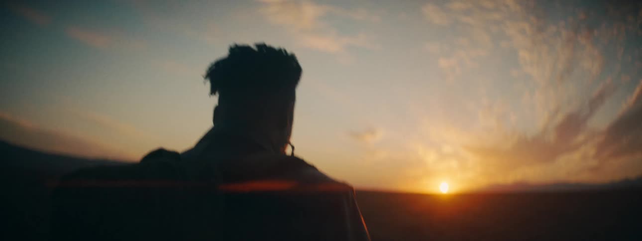 A silhouetted hiker with a backpack walks through a vast, open landscape towards the golden sunset