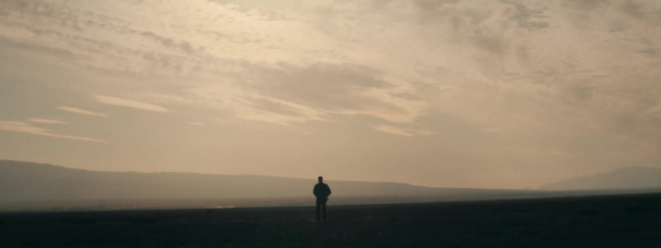 The silhouette of a man walks towards the camera across an expansive, misty plain at sunset