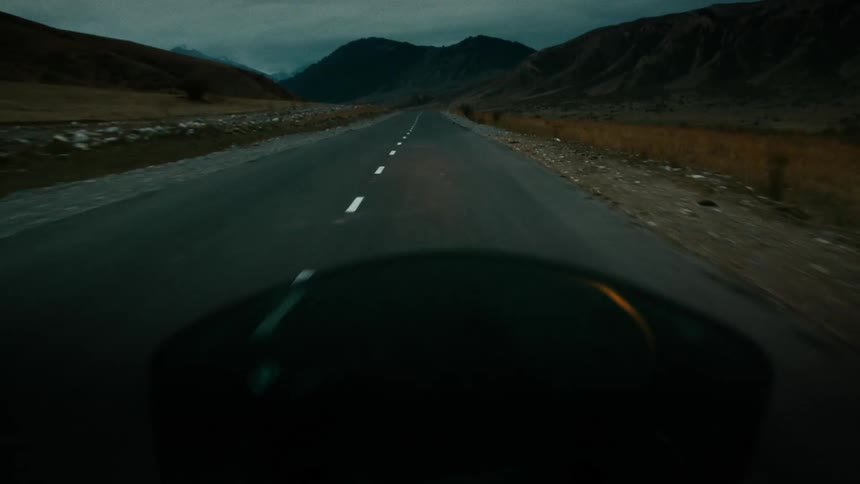A first-person view of a motorcycle driving along a dark, empty road towards the mountains at dusk