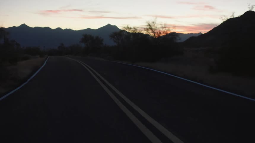 A serene drive along a winding two-lane desert road as the sun sets behind the silhouetted mountains