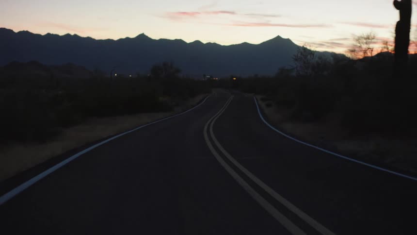 Point of view shot traveling backwards down a winding desert road at dusk, with silhouetted mountains on the horizon