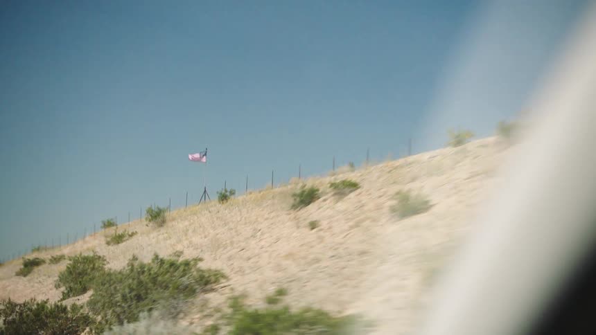 Slow motion passenger-side view of an American flag waving in the wind on a desert hillside next to a highway
