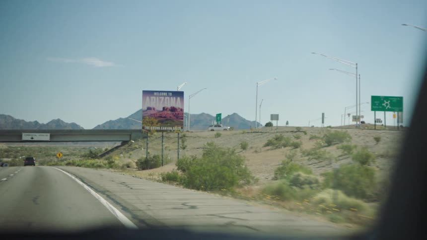 A point-of-view shot from a car driving on a desert highway approaching a 'Welcome to Arizona' sign with mountains in the background