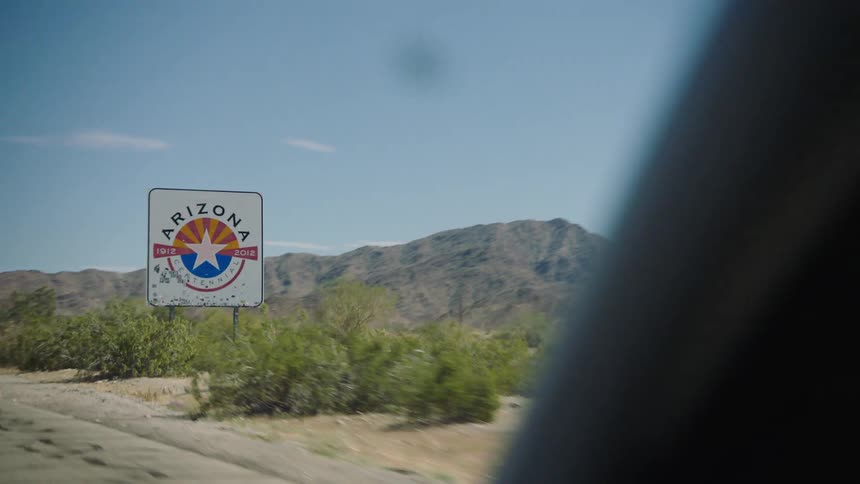 Driving past the 'Welcome to Arizona' state sign on a desert highway with mountains in the background