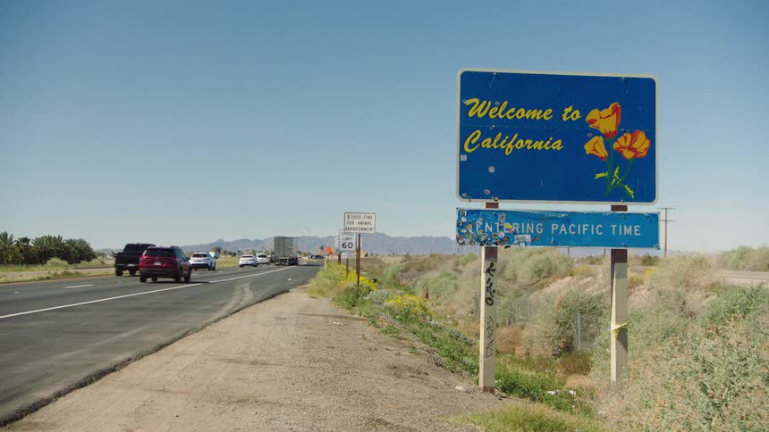 The iconic 'Welcome to California' sign stands by a busy desert highway as cars and trucks enter the state