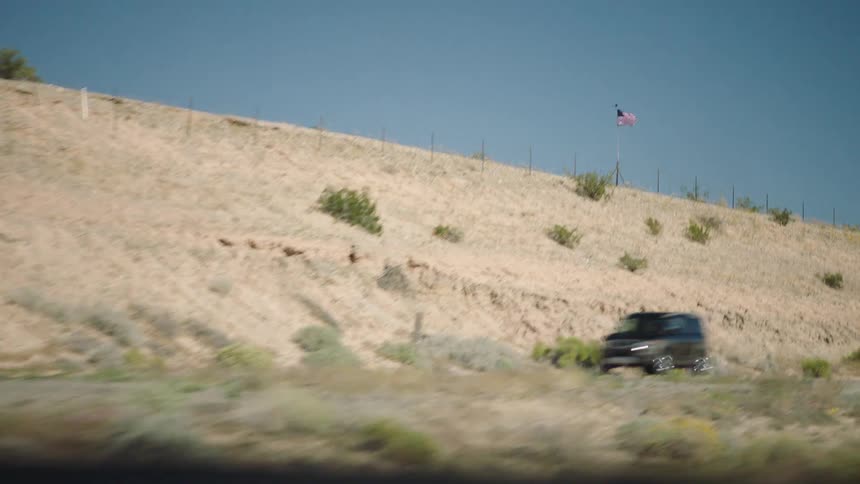 Slow-motion passenger view of an American flag waving on a desert hillside next to a highway