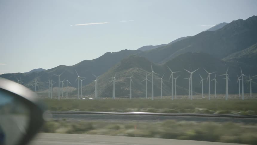 Driving past a sprawling wind farm with spinning turbines set against a desert mountain range, as seen from a car window