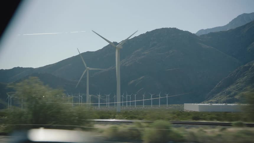 A view from a car window driving past a roadside billboard and a vast wind farm at the base of arid mountains