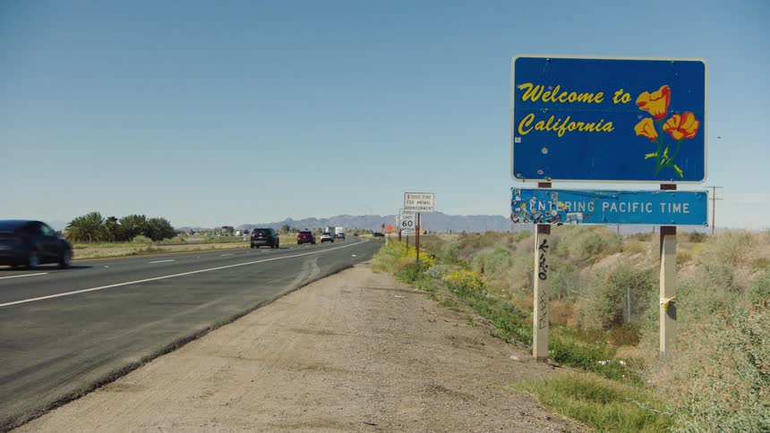 A pan reveals a 'Welcome to California' sign as traffic flows along a desert highway on a sunny day