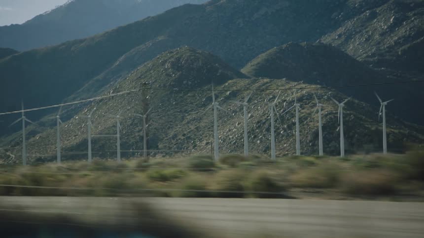 A passenger's view from a moving car of a wind farm with spinning turbines at the base of a desert mountain range