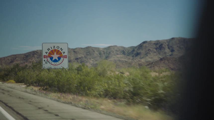 Driving on a desert highway and passing the 'Welcome to Arizona' state line sign with mountains in the background