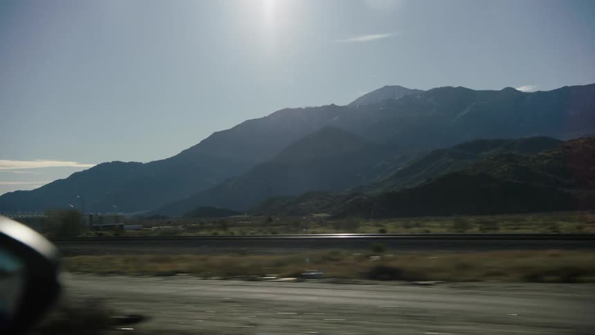 A scenic view of snow-capped mountains from a moving car driving down a desert highway next to railroad tracks on a sunny day