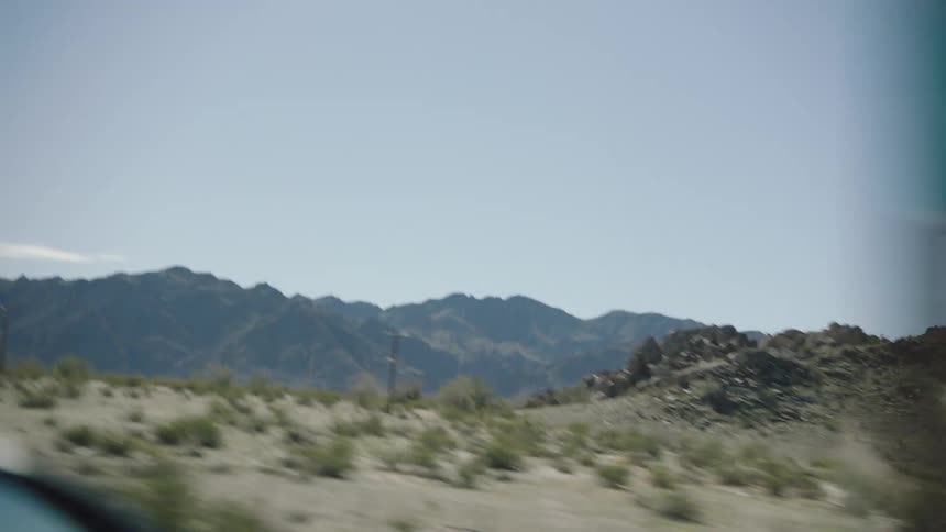 A view from a car window driving past a vast, arid desert landscape with a rugged mountain range in the background on a sunny day