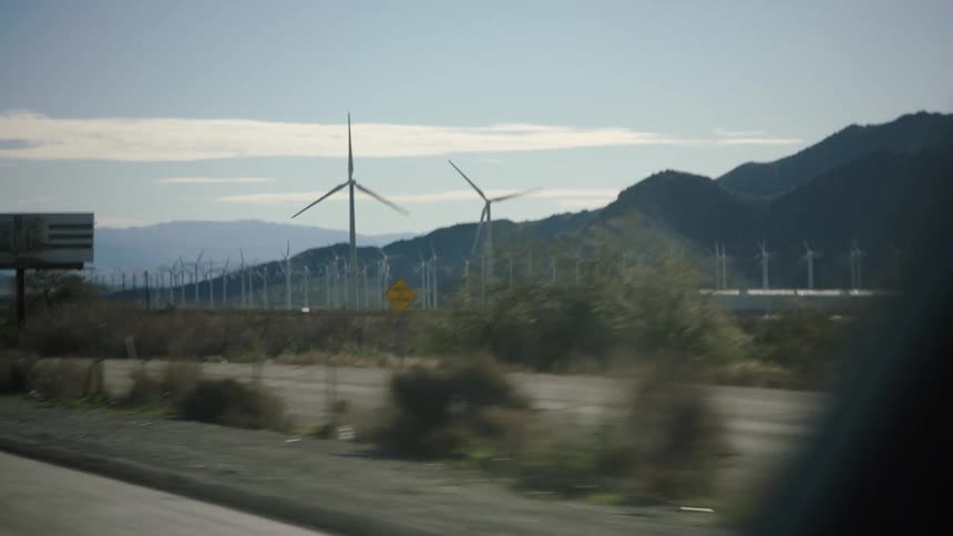 A slow-motion drive-by view of a vast wind farm with spinning turbines in a sunny desert landscape with mountains