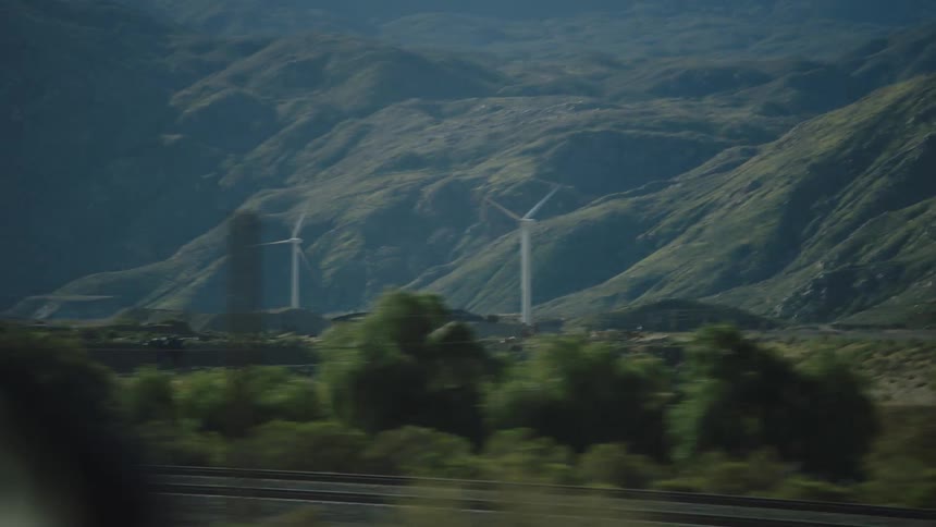 Driving past distant wind turbines spinning in front of rolling green mountains on a sunny day