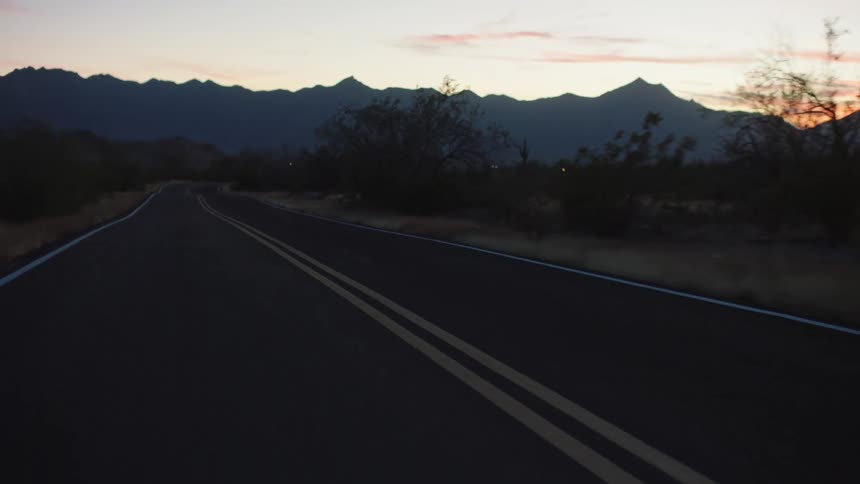 A smooth tracking shot follows an empty desert road winding towards a mountain range silhouetted against the beautiful dusk sky