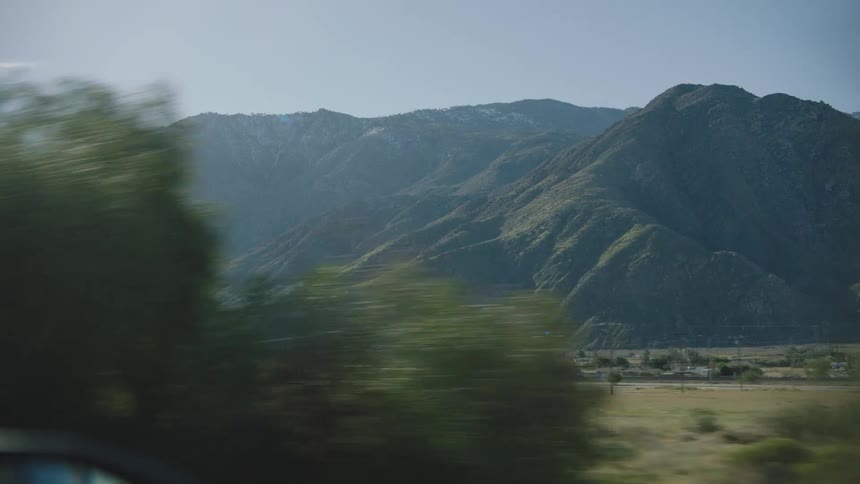 A scenic drive through a rural landscape with sunlit green mountains and snow-capped peaks in the distance, viewed from a car window