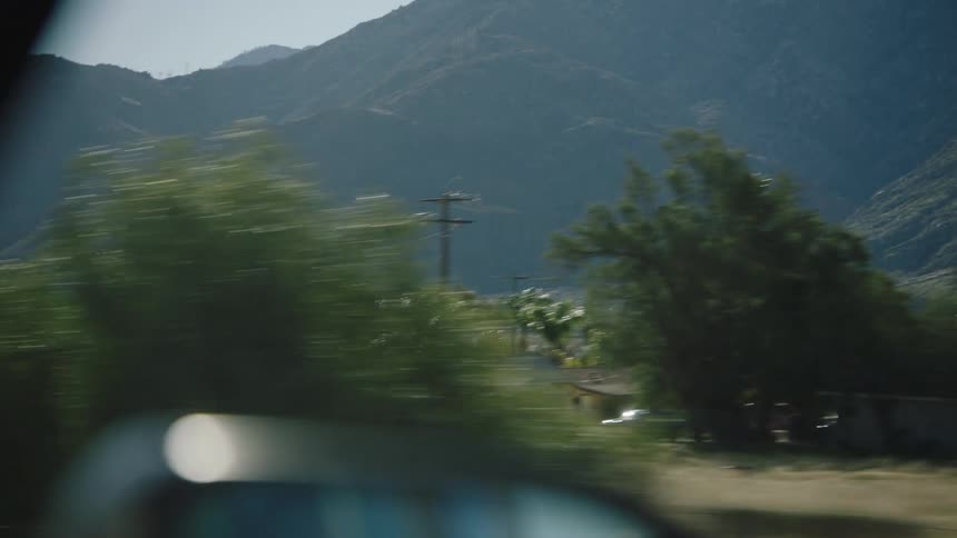 A scenic view from a car window driving through a sunlit valley with mountains and palm trees in the background