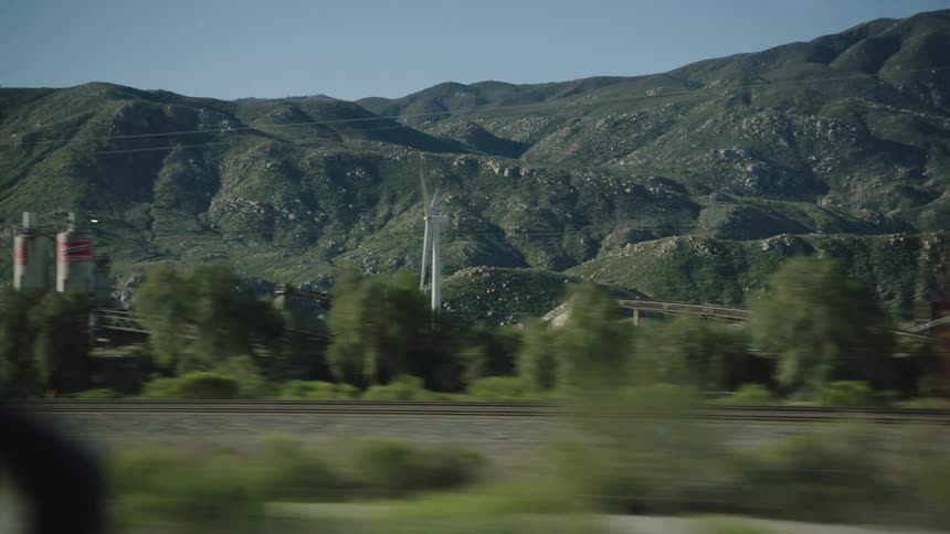 A view from a moving car captures an industrial landscape with a mining conveyor, train tracks, and a wind turbine set against rolling green mountains