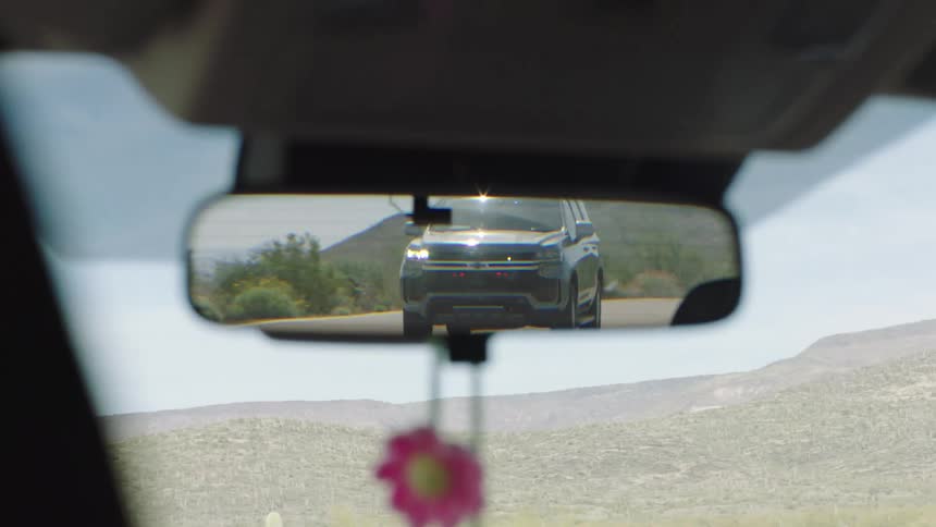 A police SUV with flashing lights is seen approaching in the rearview mirror before pulling the car over on a desert road