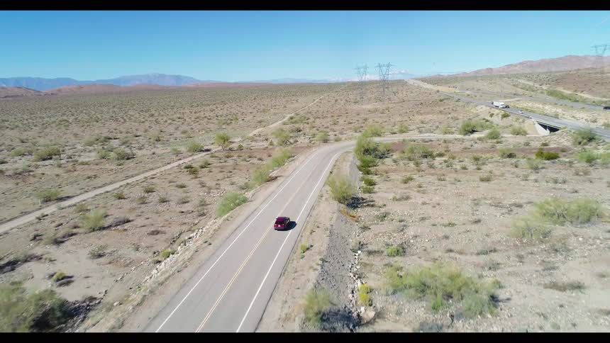 An aerial view follows a red SUV driving along a remote, winding road through the vast, arid desert
