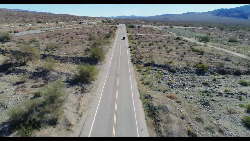 Drone footage captures a scene of stopped vehicles, including a moving truck, on a desolate desert road adjacent to a busy interstate highway