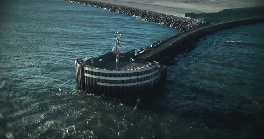 Static high-angle shot of a circular pier head and a long rocky breakwater in the choppy ocean