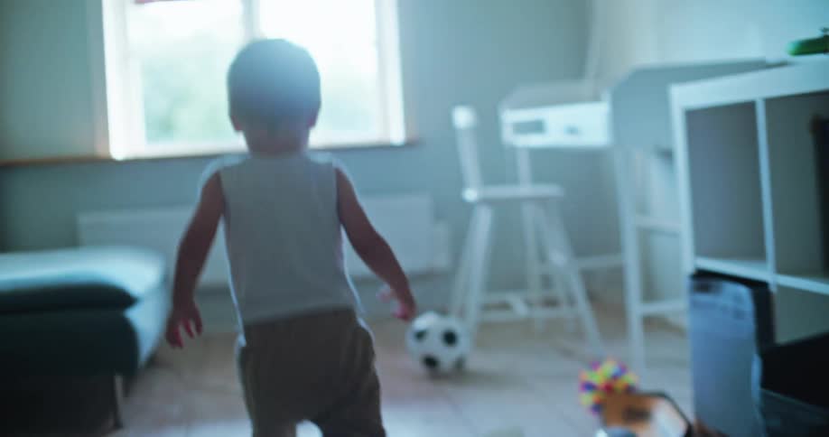 Gentle slow motion footage follows a toddler from behind as he explores his naturally lit playroom