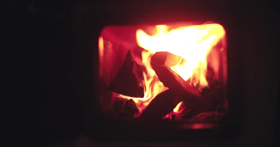 A slow, close-up reveal shows logs burning brightly inside a wood stove in a dark room