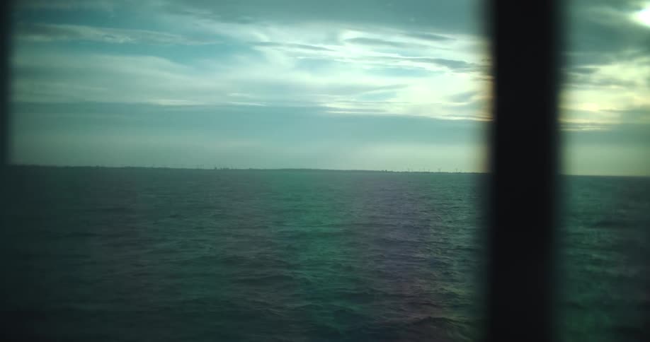 A dreamy view from a boat's window looks out over the open ocean towards a distant offshore wind farm on the horizon