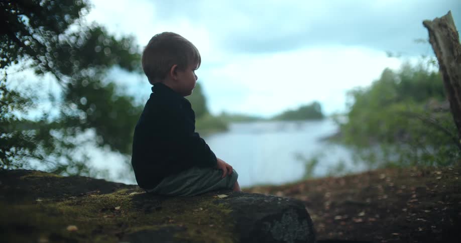 A pensive young boy sits quietly on a mossy rock, looking out over a calm lake in the woods