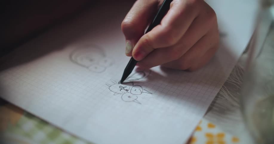 Close-up of a child's hand carefully drawing a cartoon character with a pen on white grid paper