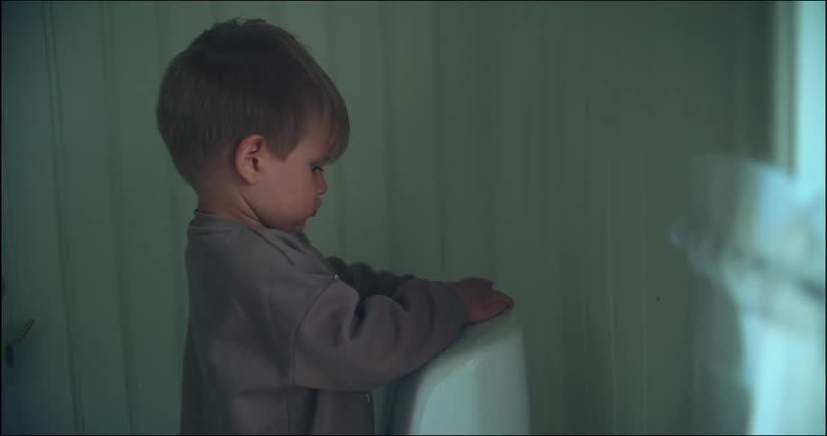 A curious toddler in brown pajamas repeatedly pushes the flush button on a toilet in a bathroom