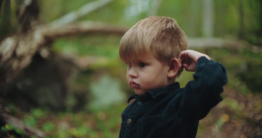 Slow motion close up of a thoughtful toddler boy scratching his head in a lush green forest