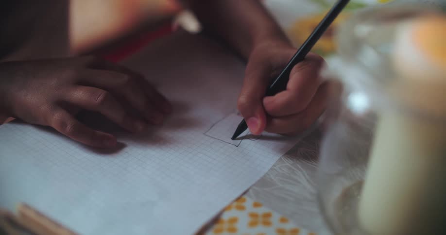 Close-up of a child's hands meticulously drawing a character on gridded paper by the warm glow of a candle