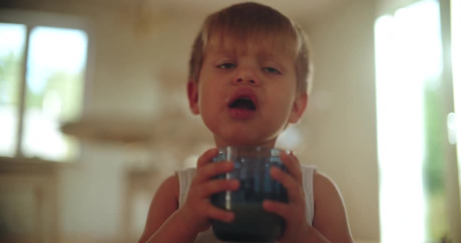 Adorable toddler boy with blonde hair drinks a beverage from a blue glass in slow motion