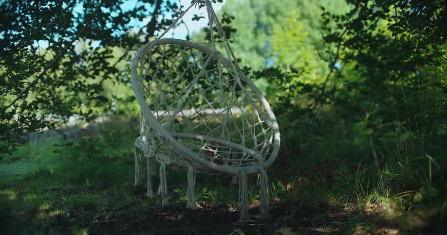 A serene white macrame swing chair gently sways in the shade of a lush green tree in a garden